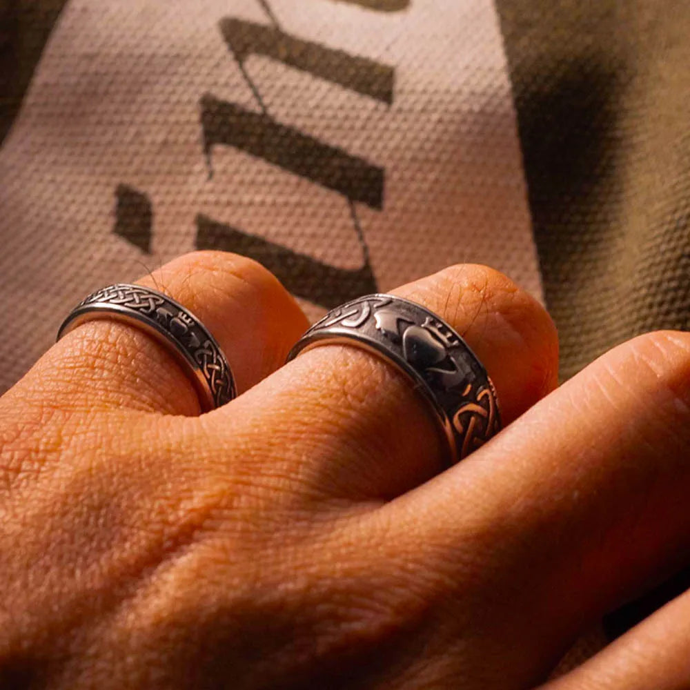 Deux bagues Claddagh en acier inoxydable sur une main, symbolisant l'amour et l'engagement, sur fond texturé.