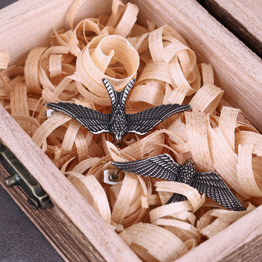 Boucle d'oreille corbeau en acier inoxydable vintage, présentée dans une boîte en bois avec copeaux de bois.